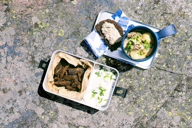 Rantakalliolla piknikastioita joissa kalaa ja dippikastiketta, keittoa ja leipää