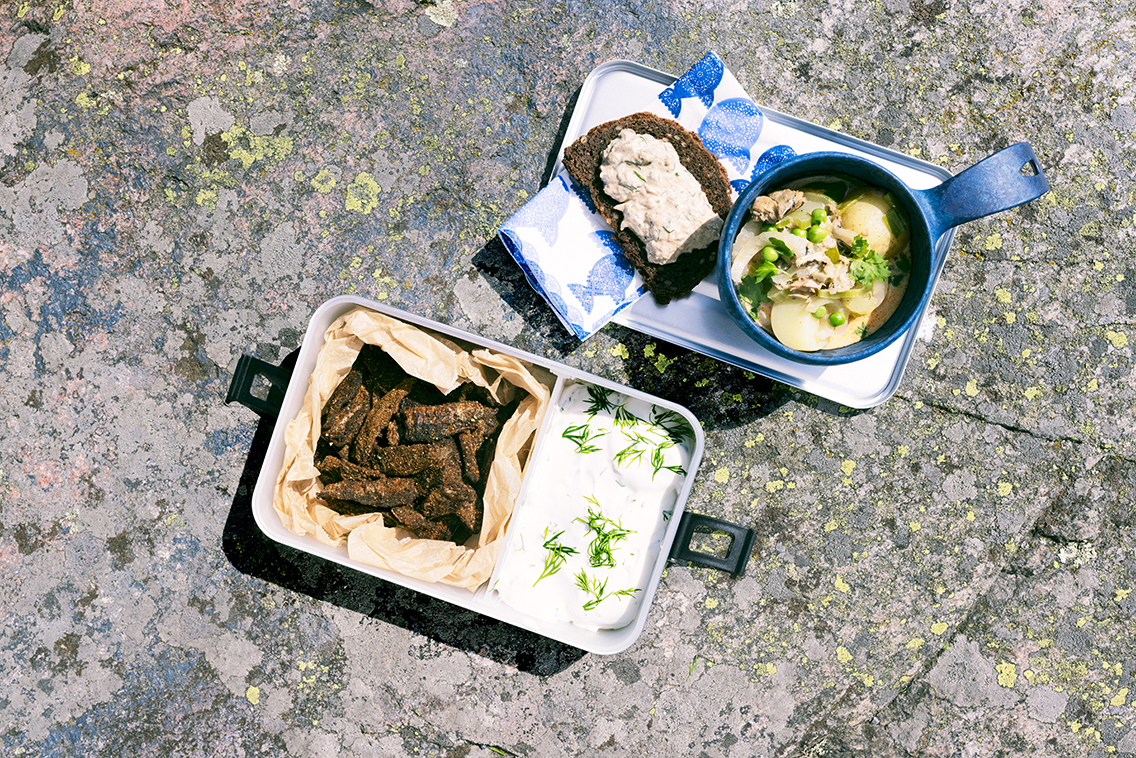 Rantakalliolla piknikastioita joissa kalaa ja dippikastiketta, keittoa ja leipää